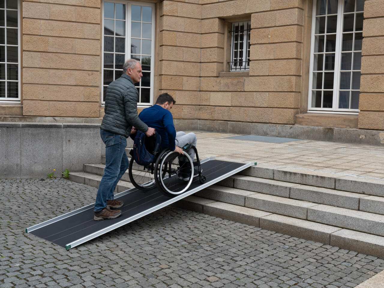 rollstuhlfahrer mit hilfsperson faehrt ueber treppe und absatz eines gebaeudes